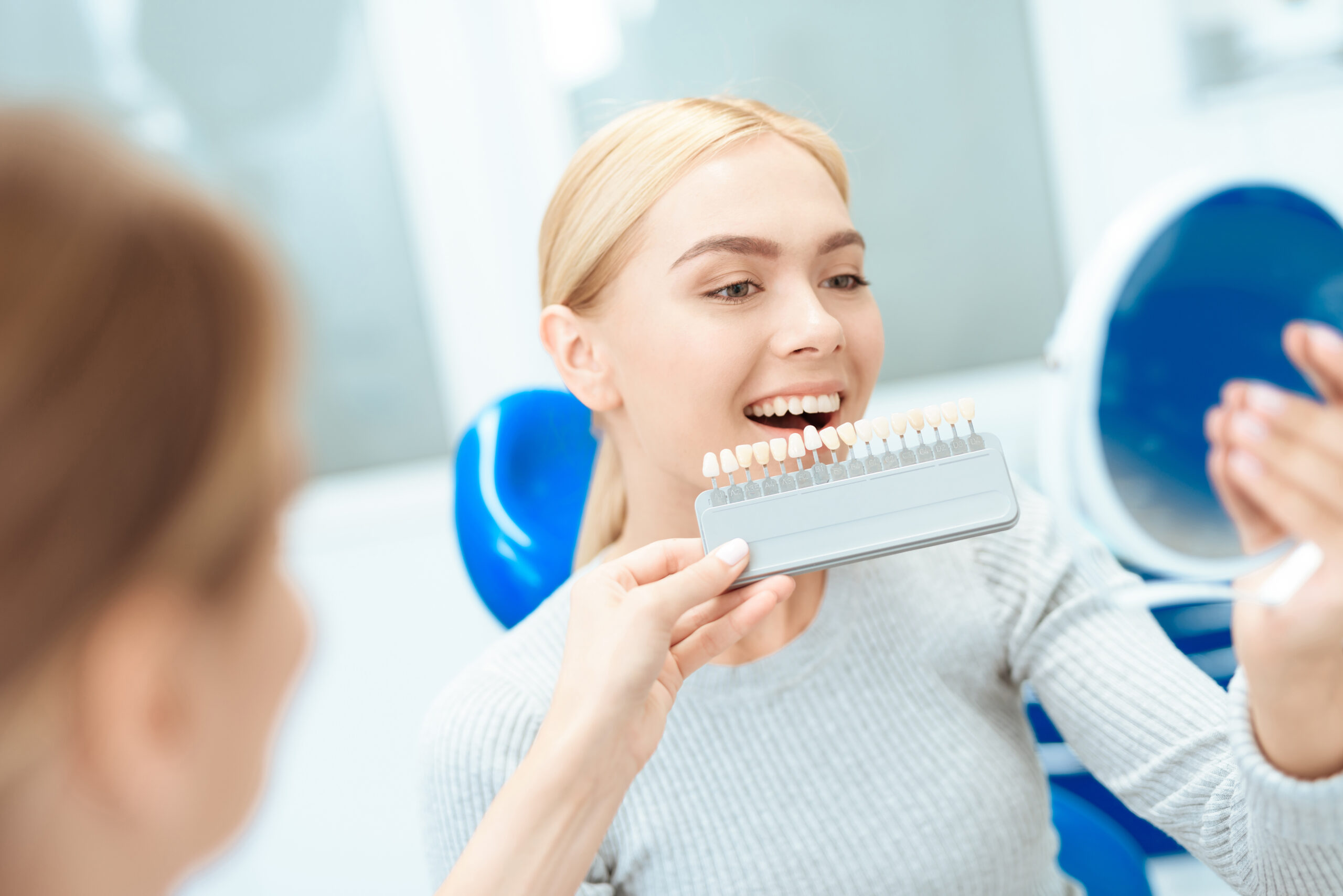 A female dentist determines the color of the patient's teeth. She holds a tooth color scale in front of the patient